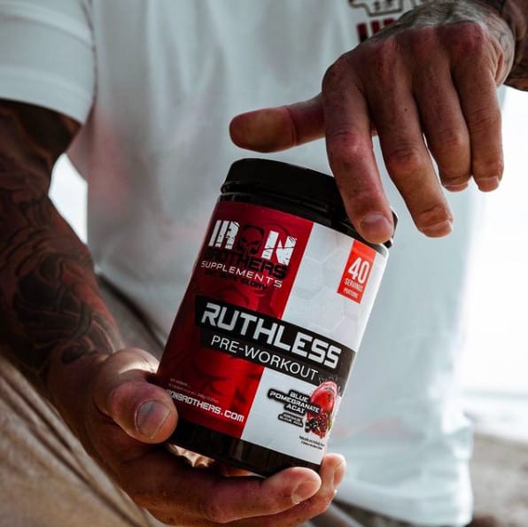 Tattooed man holding a tub of Iron Brotherhood work out supplements.